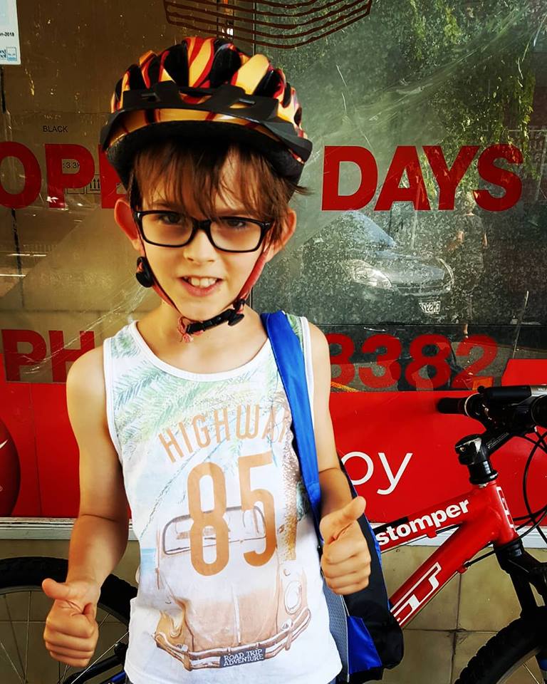 A young boy with glasses and a cycle helmet outside a bakery. He's doing the thumbs up.